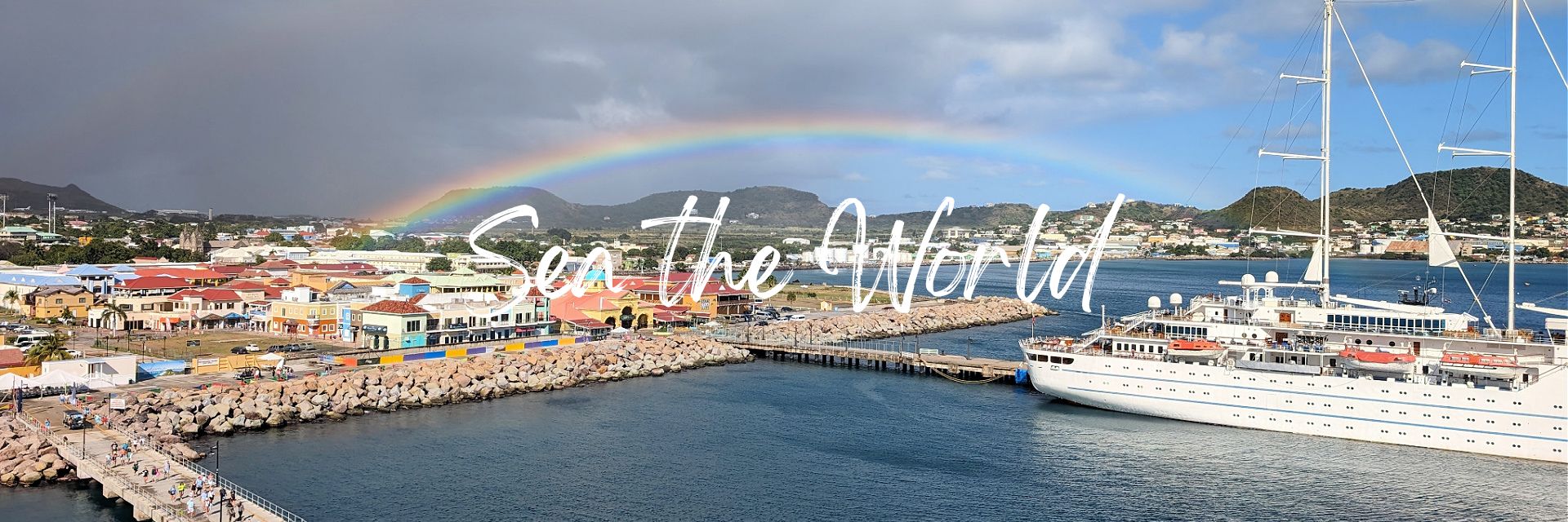 Photo of a sailing ship in the port of St Kitts. There are colourful shops on shore with a rainbow stretching across the sky. The text on the photo reads "Sea the World". 